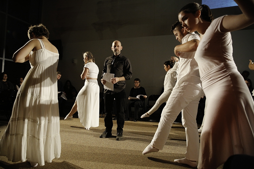 A single man dressed in black stands in the middle of five dancers dressed in white surrounding him. 
