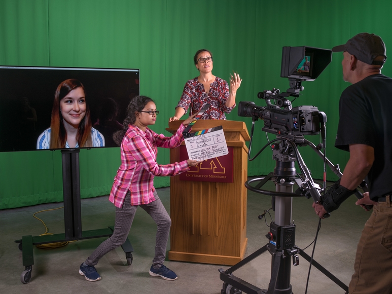 A faculty member speaks in front of a green screen while be filmed, a professional is on a video screen and a graduate student manages the room.