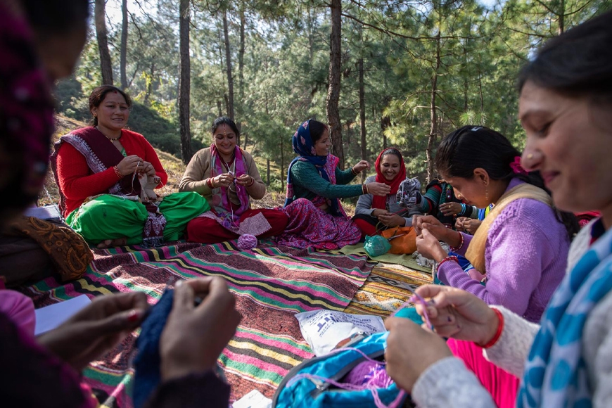 A circle of women knitting in a forest sitting on a brightly colored cloth groundcover