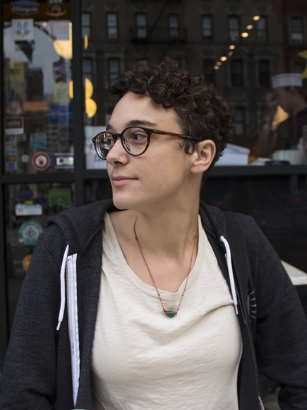 A young woman with short, brown, curly hair and glasses wearing a white tee shirt and a black sweatshirt sits looking to her right in front of a shop window