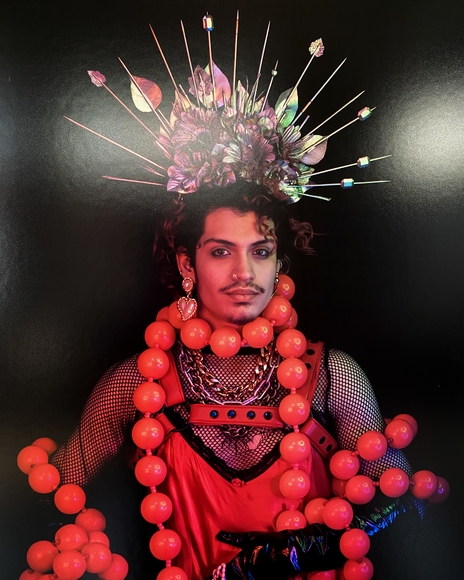 Goateed Latinx person wearing radiant spiked crown and boa of red spheres against black background