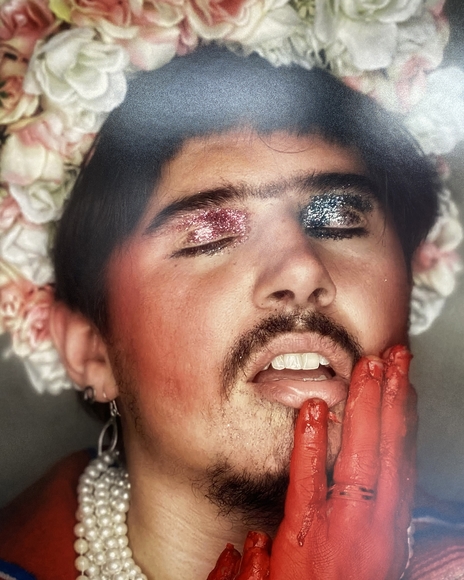 Goateed Latinx person wearing glittery eyeshadow and crown of roses touches their face with a hand painted red