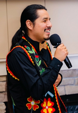 A male Indigenous student with a black ponytail and flowery vest speaks with a microphone
