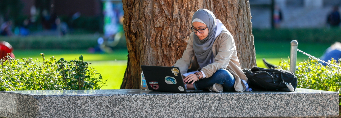 Student wearing headscarf sitting on bench working on laptop