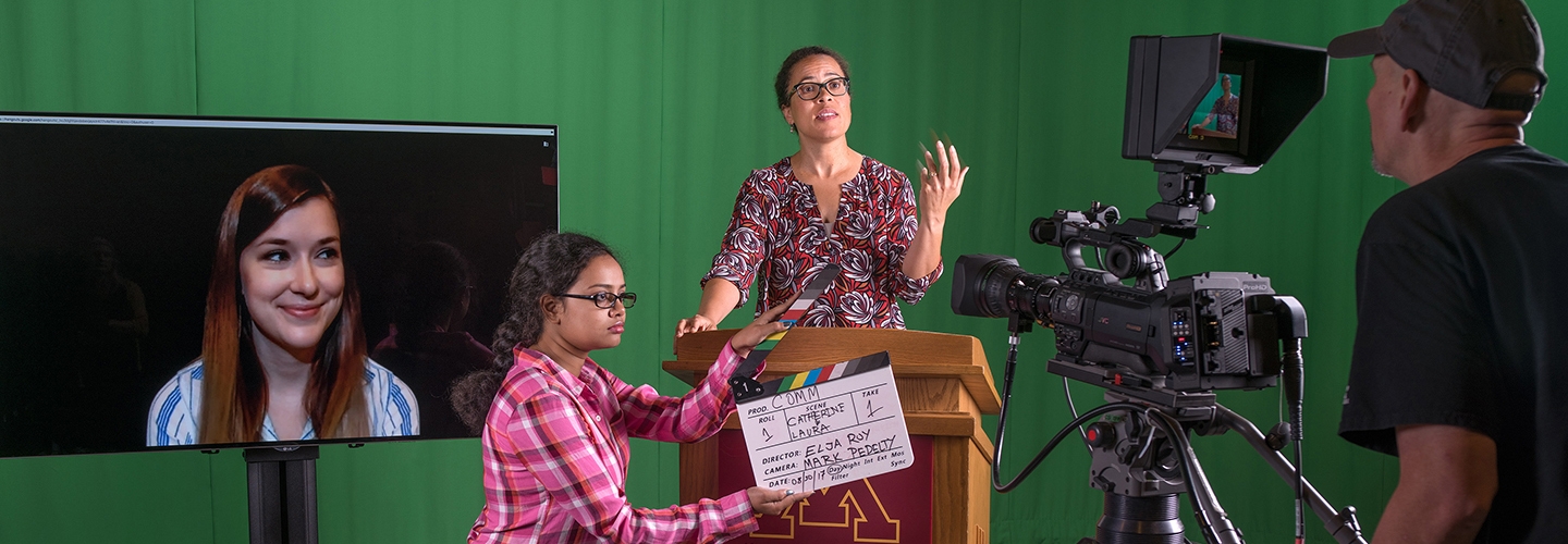 In front of a green background, a person stands at a podium. A person holds a scene marker in front of them. They both are next to a TV which is projecting the smiling face of a woman. A person records them on a big movie camera.