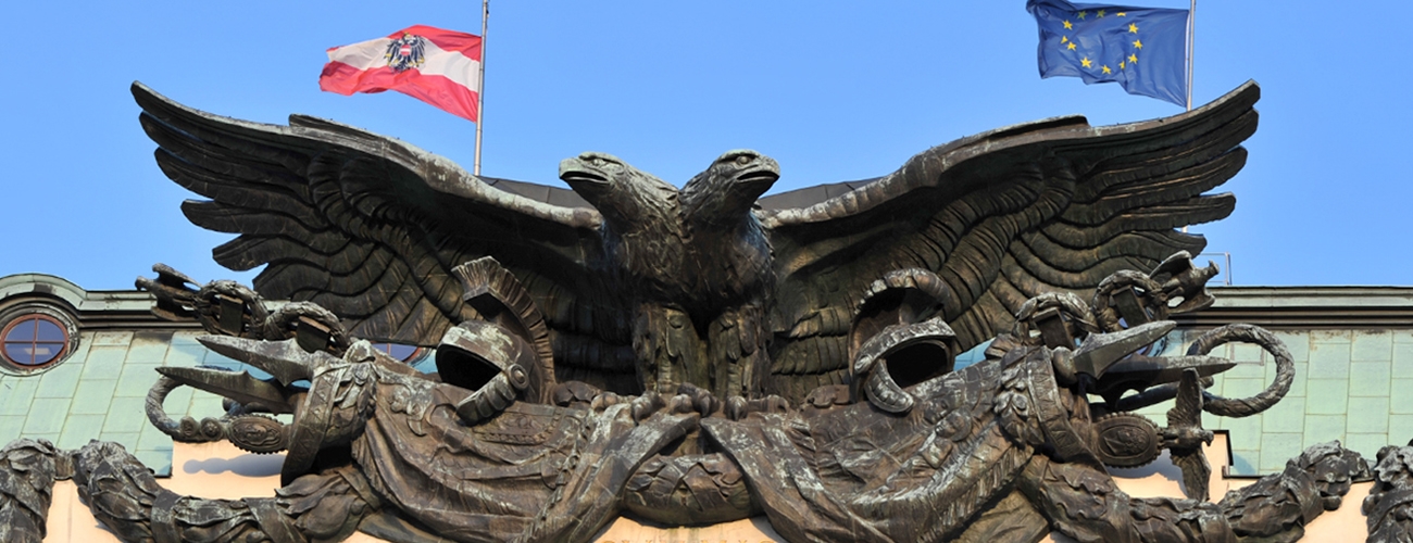 alt="Photo of the double-headed eagle on the Vienna Kriegsministerium, with Austrian and European Union flags flying behind it"