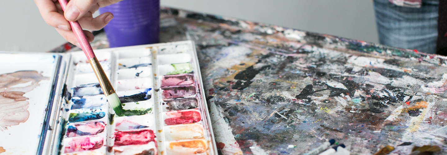 A hand dips a brush into green paint in a messy tray of watercolor paints sitting on a gray, paint-splattered table next to a pile of brushes.