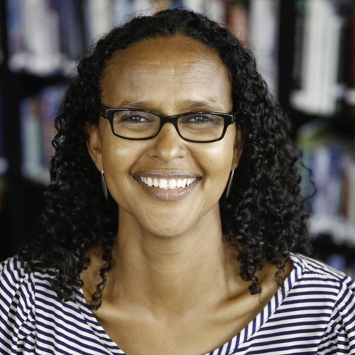 Professor Cawo Abdi, a person with shoulder length, wavy hair, light brown skin, and glasses wearing a striped top and smiling