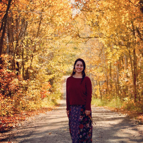 A woman with brown hair stands on a dirt road in front of yellow foliage