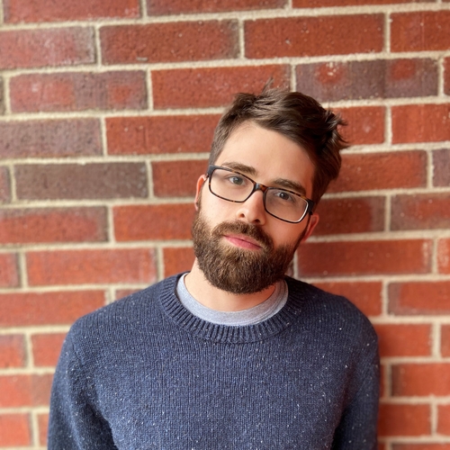 A man with glasses, brown hair, and a beard standing in front of a brick wall.