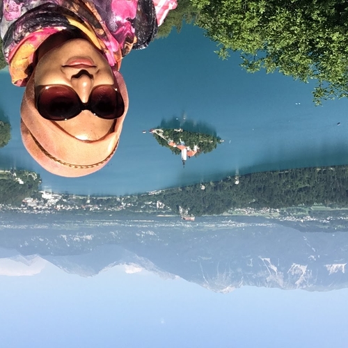smiling woman with a peach printed headscarf and sunglasses with a lake, trees, and mountains in the background