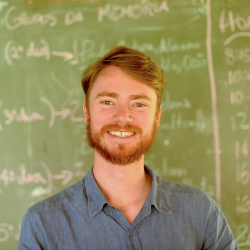Gabe Schwarzman, a person with red hair, beard, and mustache standing in front of a chalkboard wearing a blue buttoned shirt