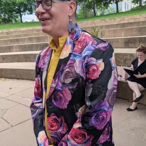 A person wearing a colorful floral jacket and earrings; they are smiling.
