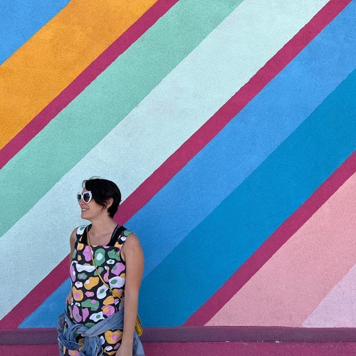 Woman in multicolored tank top & sunglasses stands in front of colorful striped wall