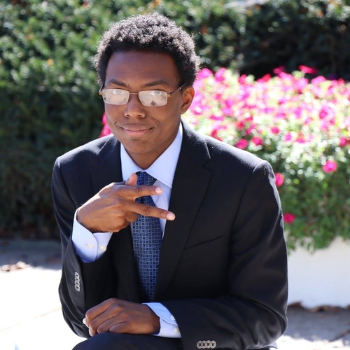 An image of A Adams, a black non-binary masc person with a short Afro. They are wearing a black suit, powder blue button-up shirt, a glossed Navy tie, and glasses. They are flashing two fingers, like the peace hand sign, sideways.