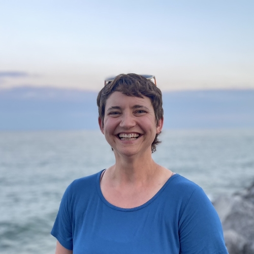 A white woman with short brown hair and a blue shirt looks straight at the camera with a big smile. Behind her is a background of blue water, a sunset with light pink and blue clouds, and large gray rocks. Photo Credit: Jamie Ryan-Karels