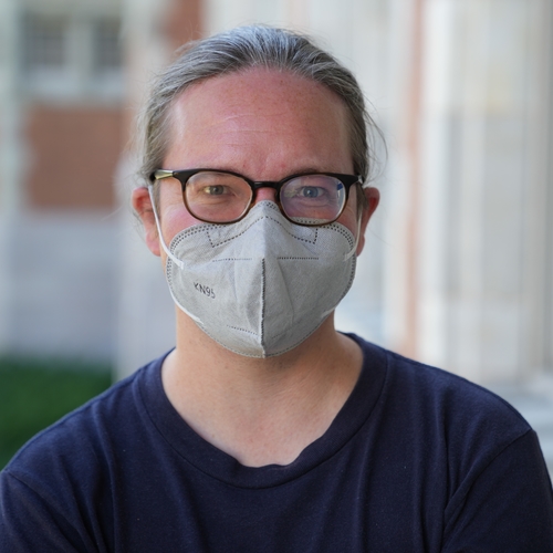 Person wearing glasses and a mask with Folwell Hall in the background