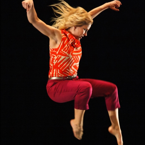 Laura Selle Virtucio, with long blond hair, wearing red pants and an orange shirt, is picture mid-jump.