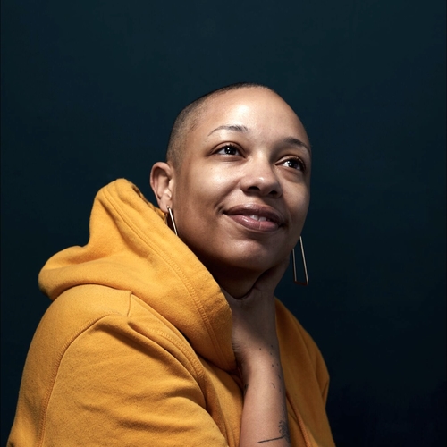 a light skinned black woman with a shaved head looks up to her right. she is wearing a yellow sweatshirt and her right forearm is across her chest and her hand rests on her neck.