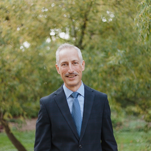 A 48 year old grey haired white man is seen centered, in a blue suit and tie. He stands in a tree filled garden.  