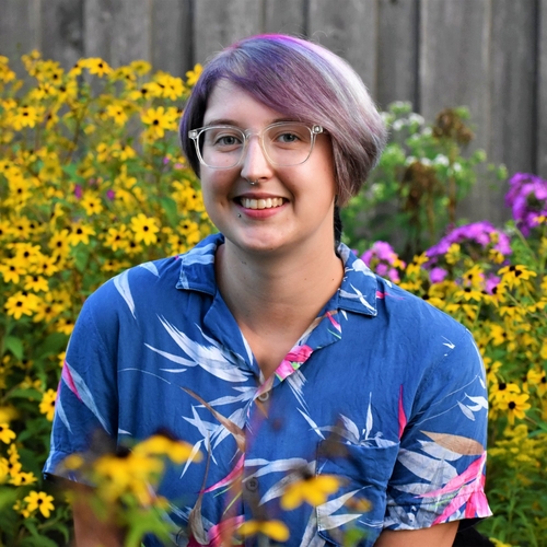Korrina is smiling. She has grey, pink, and white hair and is wearing a blue shirt. They are surrounded by yellow and purple flowers