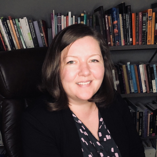 2022 Profile picture of Jessica A. Cooley. Image ID: White woman with shoulder length brown hair, smiling and wearing a black blazer and purple dotted blouse, and seated in front of her bookshelves. Looking pretty pro, kinda shiny, and generally like "hey what's your favorite book?" woo woo!