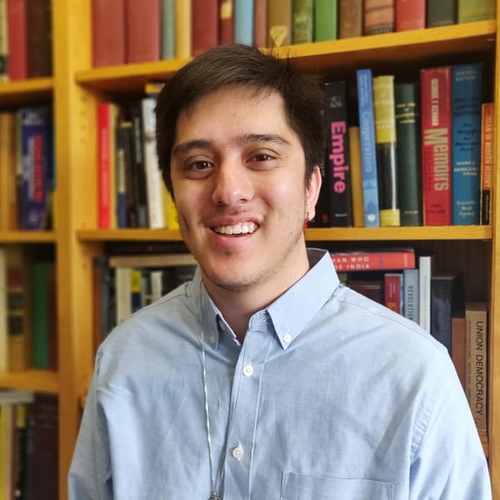 A picture of Adam wearing a blue dress shirt. Adam is in front of a bookshelf and is smiling.