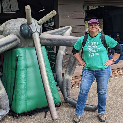 Katherine Klink, a woman with grey hair and light skin, wearing a purple cap, a green t-shirt and blue jeans, and standing next to a large inflatable mosquito.