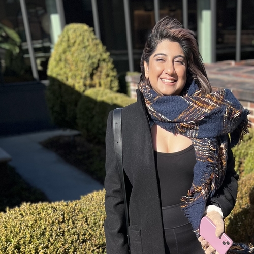 a south-asian, indian, non-binary person smilingly widely with their hair flowing on a windy rooftop balcony with blinding sunlight. the person, saumya, is wearing a black top with a black coat and a blue and brown checkered scarf. they’re wearing brown gloves with white fur and holding a lavender phone in the picture. the background features lush green bushes. 