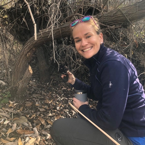 A photograph of Dr. Fairfax squatting down next to a beaver chewed stick, holding up a small stuffed animal beaver. She is wearing a purple jacket and grey pants.