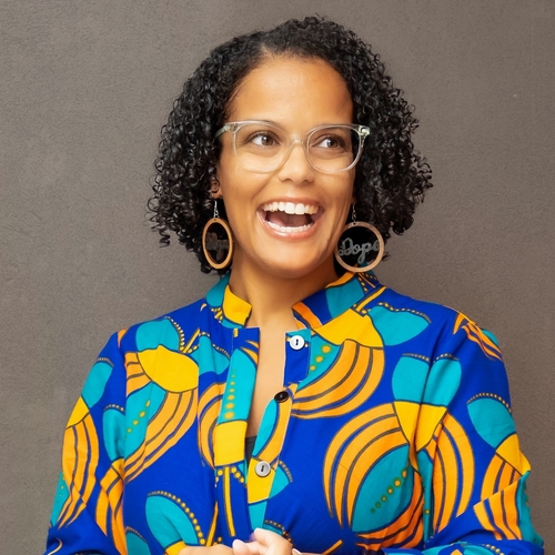 Photo of a Black woman with curly black hair, smiling, wearing glasses and a brightly colored shirt.