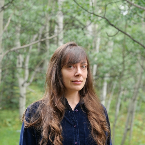 a woman with long brown hair wearing a dark blue velvet button-up shirt stand before a grove of Aspen trees