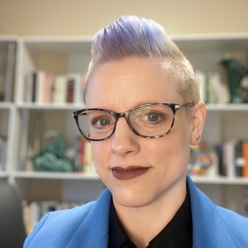 Photograph of a white woman with short purple hair, glasses, and dark lipstick wearing a black shirt and blue blazer