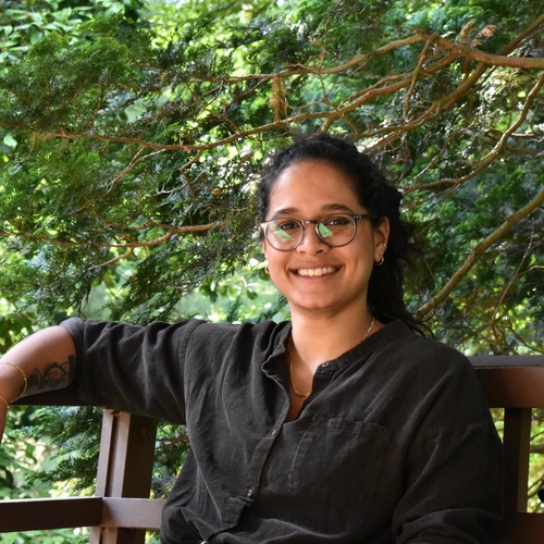 A young South Asian woman in greay, round, acrylic glasses and dark, curly brown hair in a ponytail smiles at the camera. She is seated on a bench against a backdrop of trees.