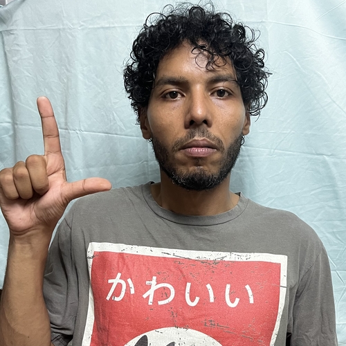 The image shows a man with curly hair and a beard, wearing a grey t-shirt with Japanese characters and an illustration on it. He is making an "L" shape with his left hand, holding it up next to his face. The background is a light blue sheet.