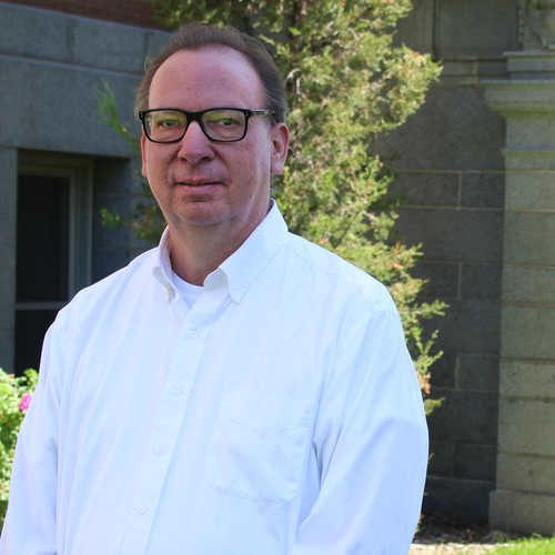 A man wearing glasses and a white button-up shirt stands outdoors in front of a stone building and greenery.