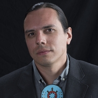 Light-skinned American Indian male in his late 30s with dark brown hair and eyes, wearing a beaded turquoise blue turtle bolo tie and a dark gray suit and light gray shirt, against a dark background
