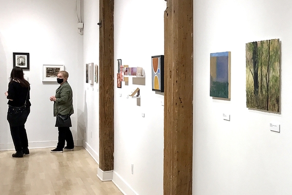 Two people talk in a gallery, surrounded by small works of art on the walls