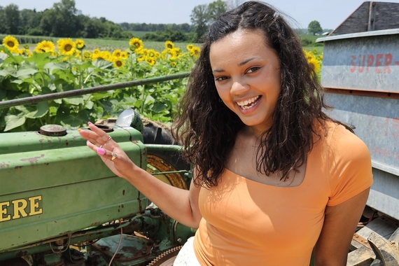 Head and torso of person with dark wavy hair to shoulders and light brown skin, smiling and wearing orange top and gestering at farm equipment and field of sunflowers