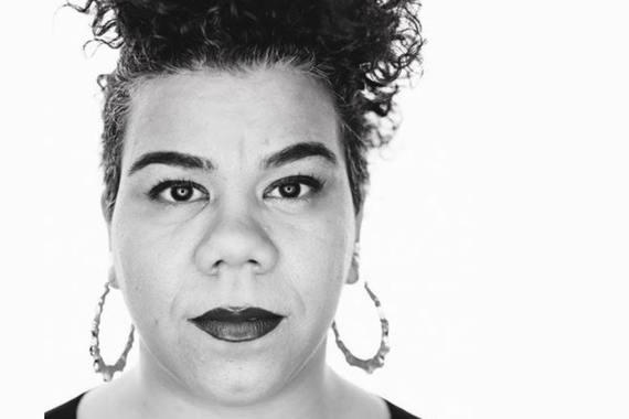 Portrait of a person looking directly in the camera with short curly hair, dark eyebrows, and dark lipstick, wearing large hoop earrings. Photo is shot in black and white.