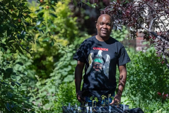 Lamar Peterson, a Black man wearing a black Cymande band shirt, stands in his backyard garden