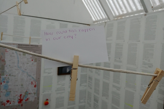 A close-up photo of a hand-written card that says "How could this happen in our city?" The card is clipped to a rope by a clothespin.