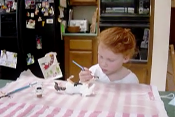 screenshot of young, red haired child painting at a table