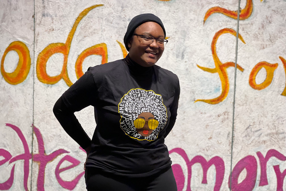 A Black woman wearing all black smiles in front of a painted plywood mural