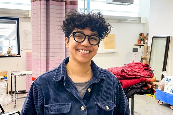 A young person in a blue corduroy shirt and black glasses smiles in a cluttered art studio with boxes of clay, piles of fabric, and a tower of pink insulation foam.