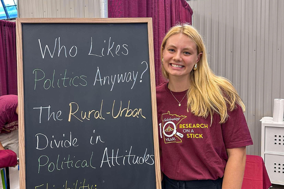 Brianna Kreft next to chalkboard reading: "Who Likes Politics Anyway? The Rural-Urban Divide in Political Attitudes."