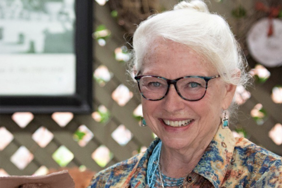 Head and shoulders of person with white hair and light skin, smiling and wearing dark rimmed glasses and patterned top; background a wood lattice with framed items