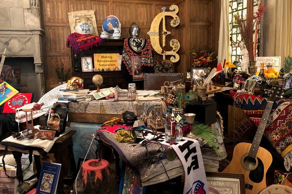 Jumble of books, artifacts, musical instruments, and textiles in a paneled room with a stone fireplace and stained glass window.