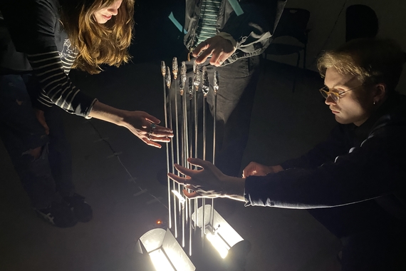 Students gently touch a gridded, vertical array of metal rods, alit from the bottom of the structure.