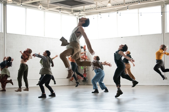 Scene from "A Dream of Touch When Touch Is Gone" featuring several dancers in neutral-colored clothing, six feet apart, in different phases of jumping: some about to take off, some just landing softly, some still mid-air. they all face away from the camera and toward a high window with light streaming in.